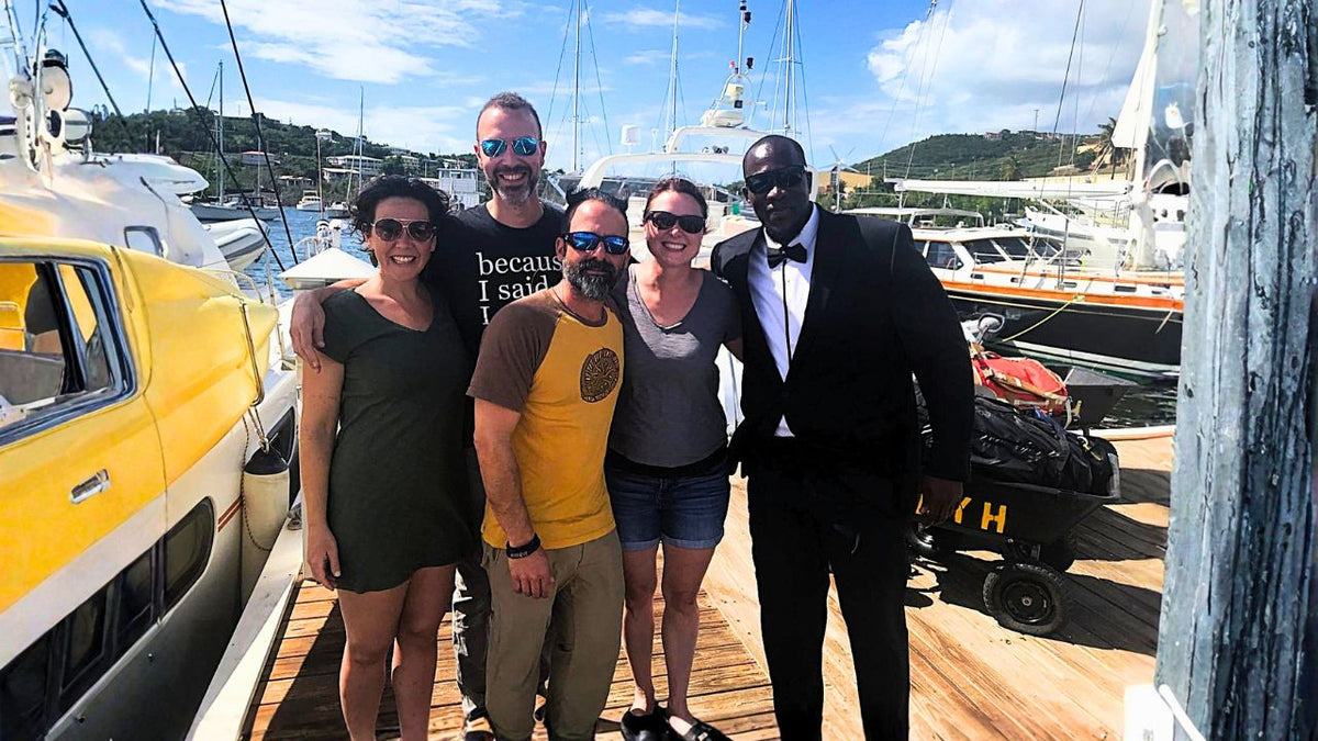 Five people posing together on a wooden dock next to a yellow boat, with yachts and hills in the background under a blue sky.