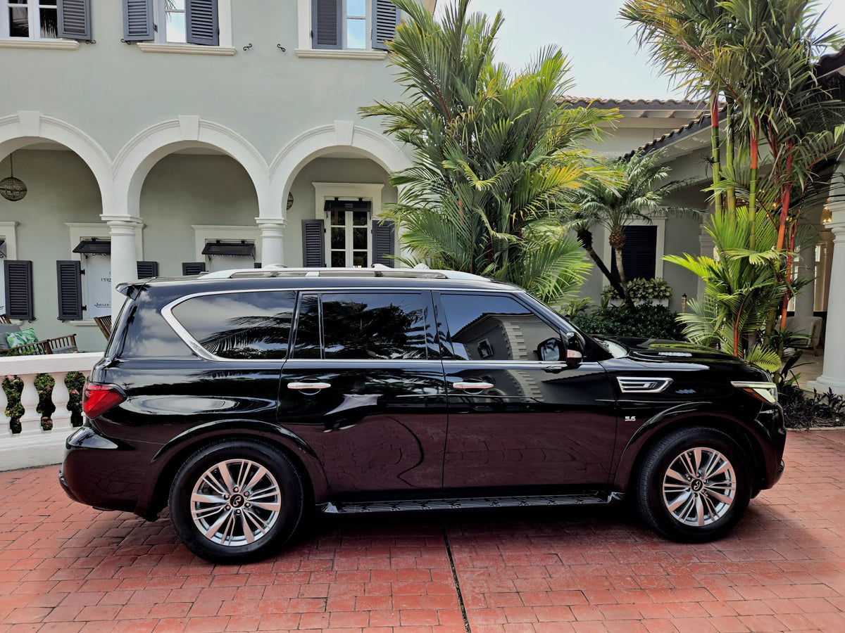 A black luxury SUV parked on a red brick driveway in front of a large white stucco house with arches and palm trees.