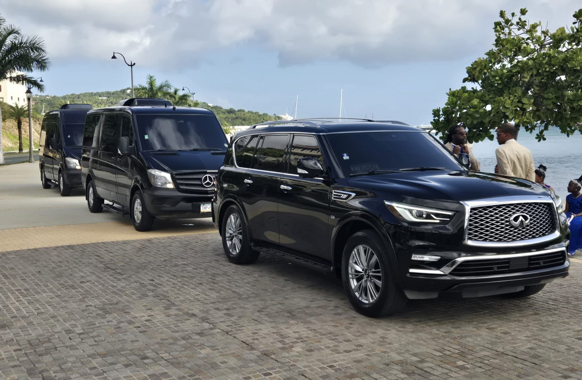 A black luxury SUV with a chrome grille and headlights is parked on a cobblestone road outdoors, with other black vans behind it and people standing near trees and water in the background.