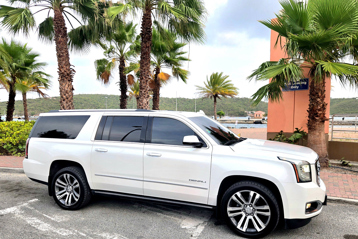 A white Cadillac Escalade SUV parked outdoors on a paved area with palm trees, a building, and water in the background.